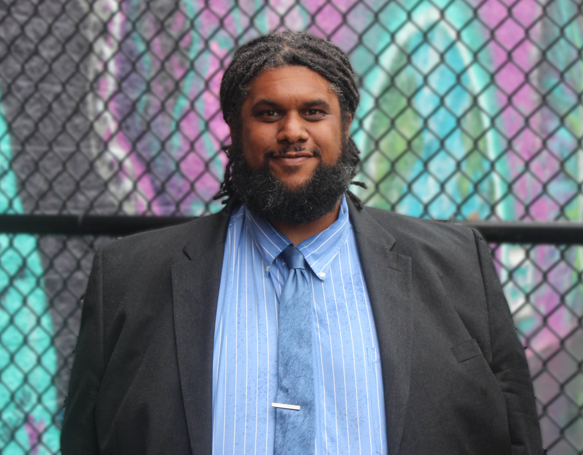 Picture of Jordan Berg Powers, a large Black man with black dreadlocks pulled back behind his head and a black beard. He is standing in front of an aqua and purple mural blurred out. Wearing a gray suit jacket, blue shirt and blue tie.